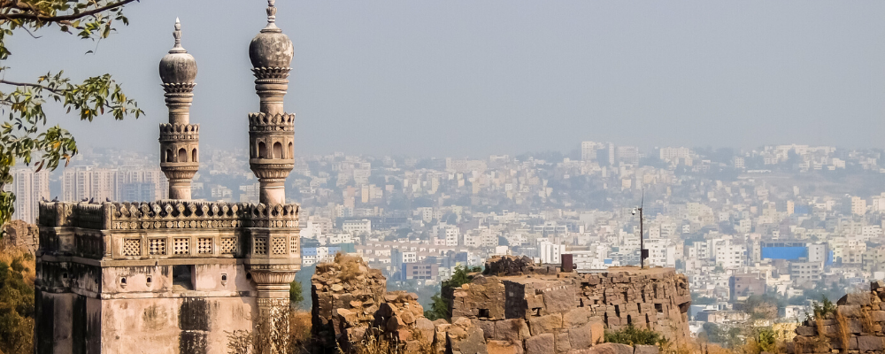 Ancient Indian fortress ruins with minarets overlooking a hazy city skyline.