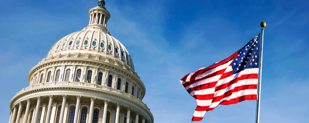 The white dome of the United States Capitol building standing next to a waving American flag against a bright blue sky.