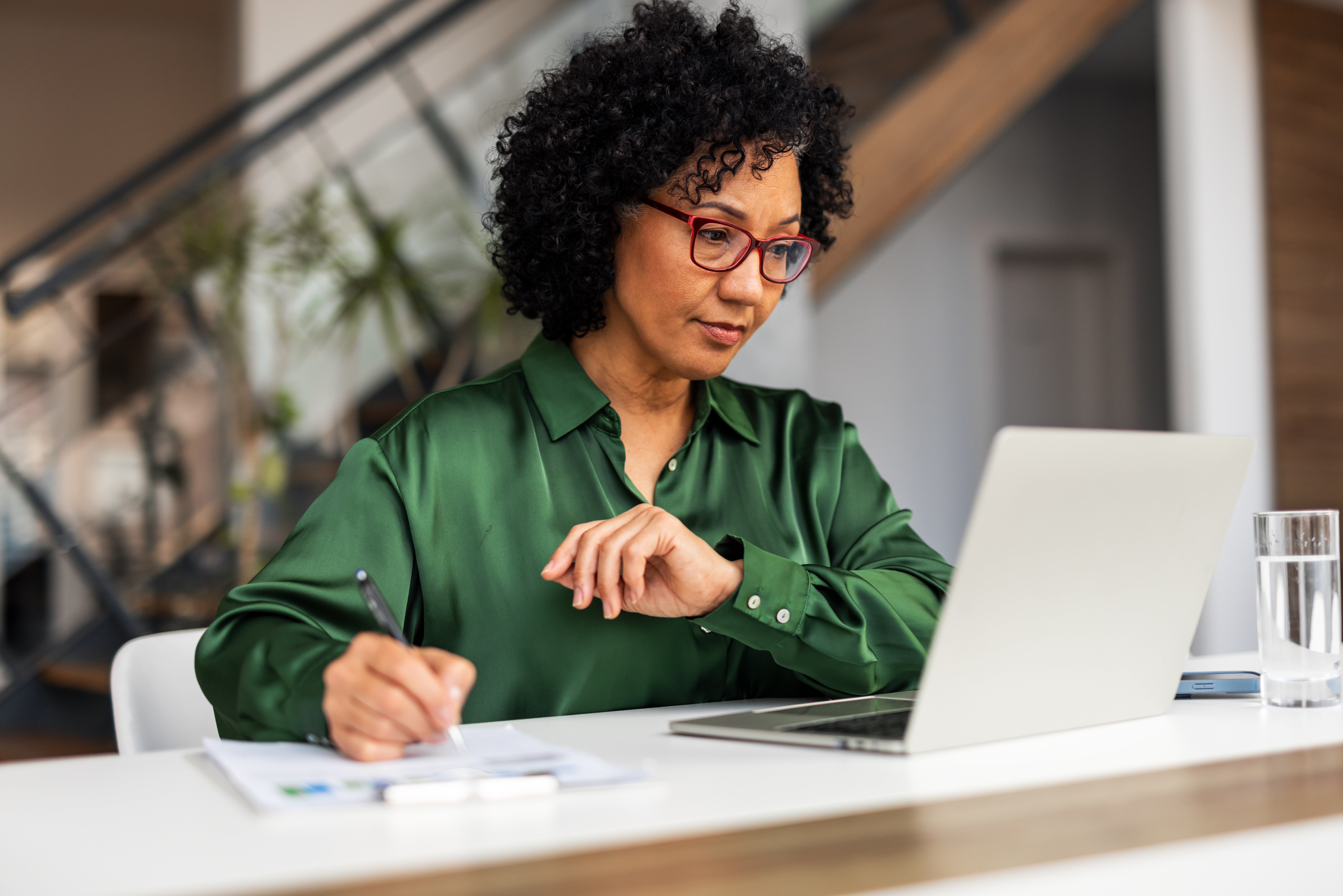 A concentrated professional woman in a green shirt works diligently on a desk project using a laptop. The setting includes bright lighting, modern design, and a productive environment.