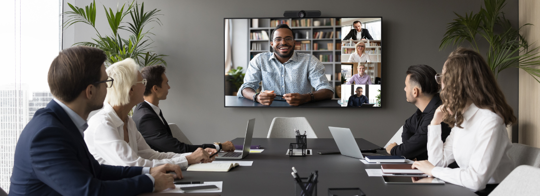 A diverse, hybrid corporate meeting illustrating global team alignment. Five professionals sit around a modern boardroom table with laptops and notebooks, looking toward a wall-mounted monitor.